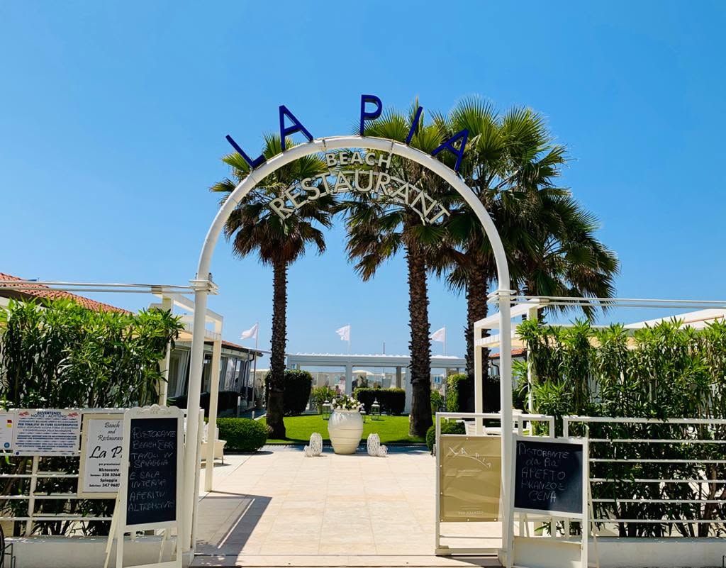 Ingresso di La Pia con arco bianco, insegna blu e palme in una giornata di sole