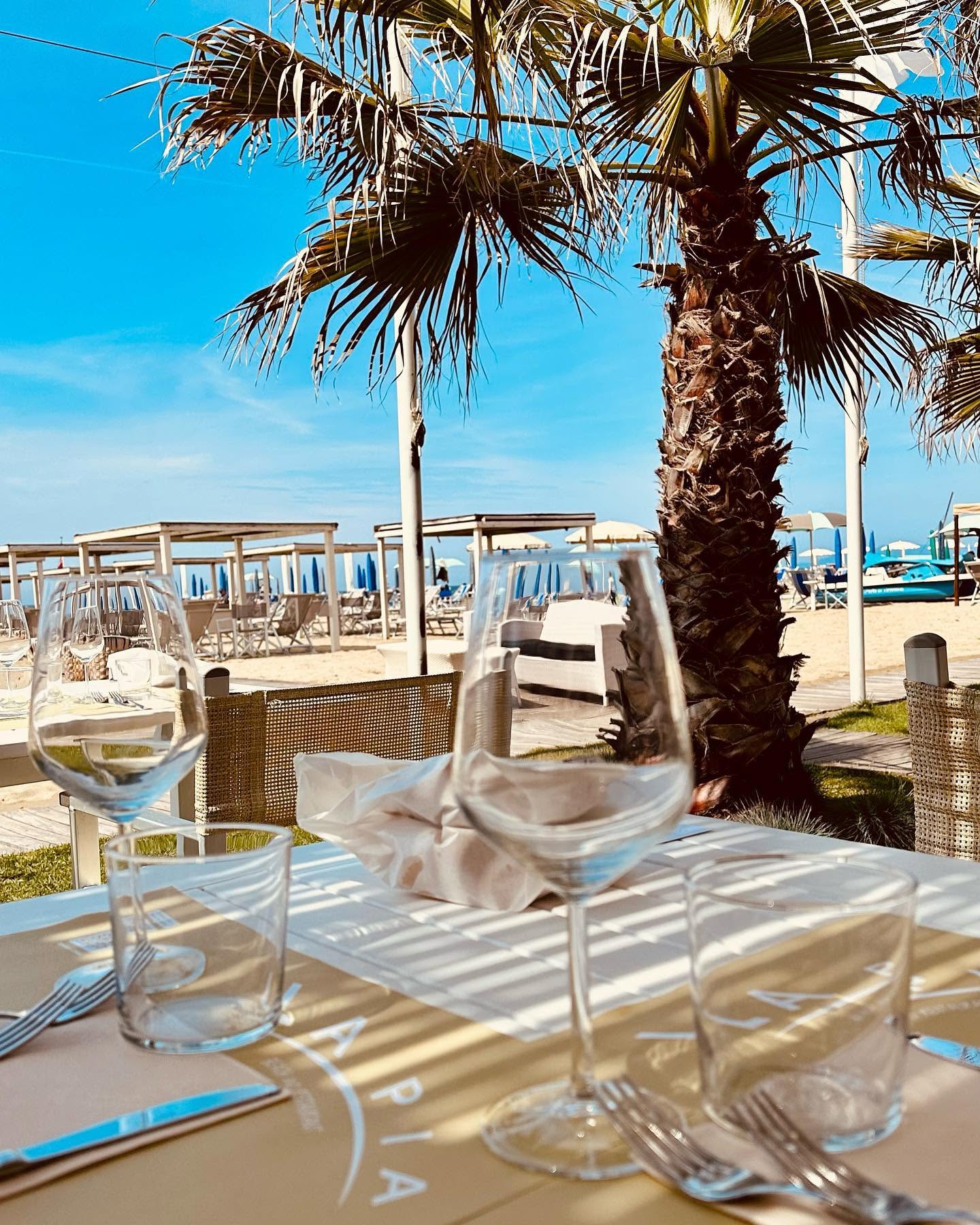 Tavolo apparecchiato sotto una palma con la spiaggia e gli ombrelloni sullo sfondo